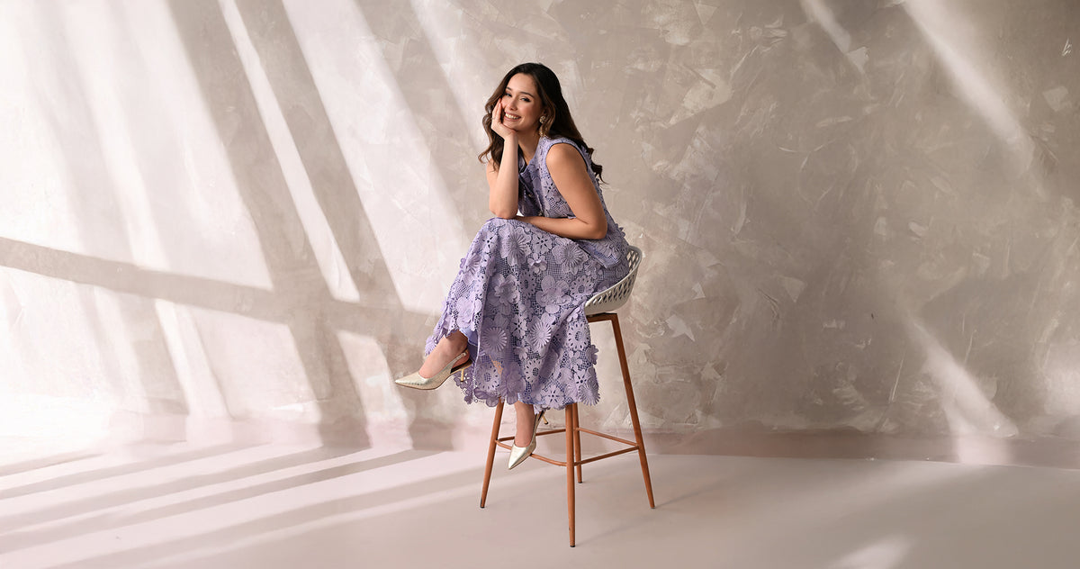A model in a lilac floral lace dress from the Beyond Sunshine collection, seated on a wooden stool against a sunlit plaster wall.