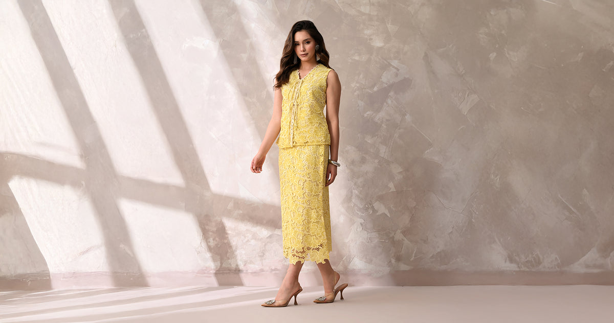 A model in a yellow floral lace top and skirt set from Anysa, standing against a sunlit plaster wall.